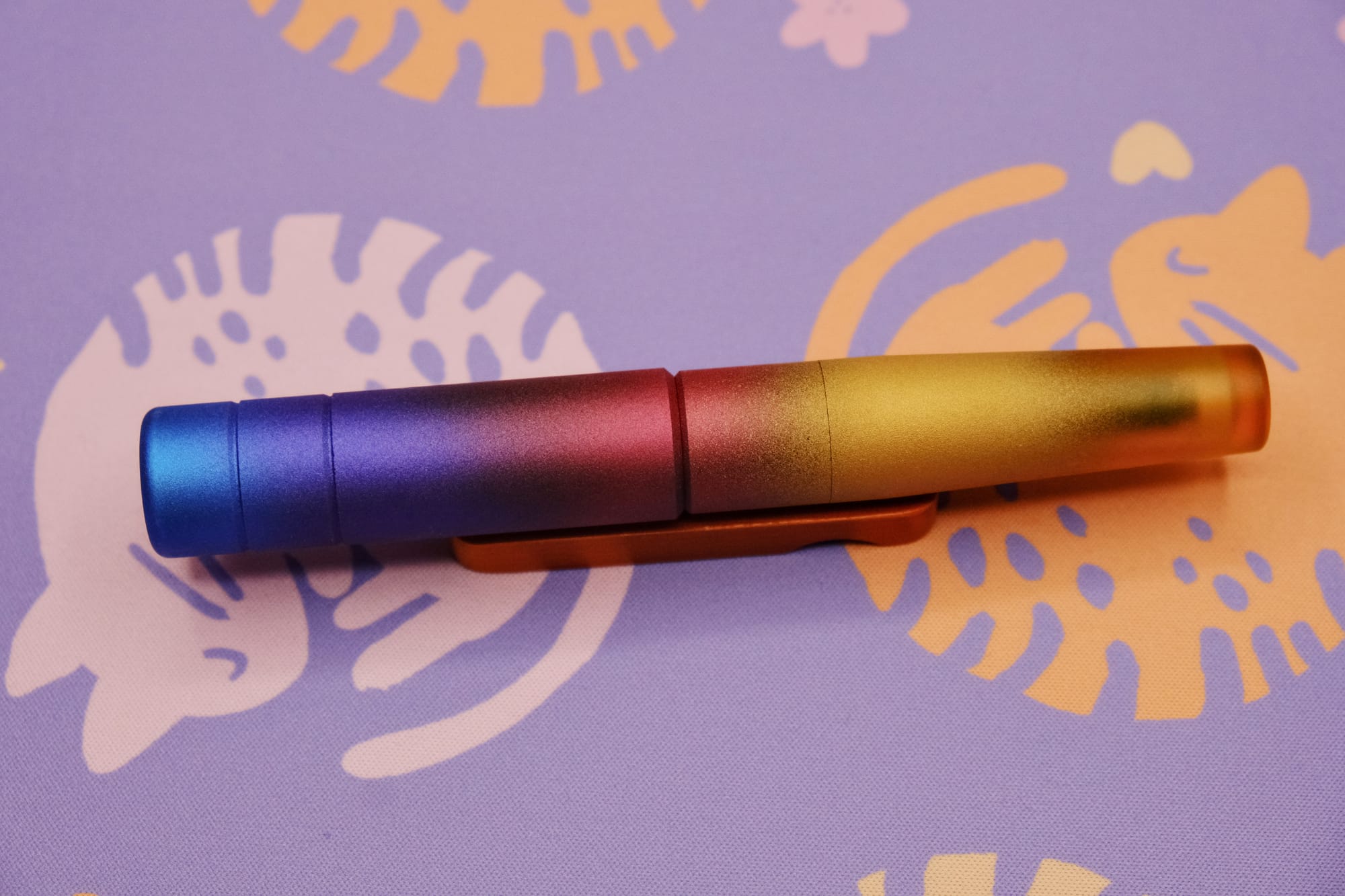 A relatively large cylindrical fountain pen with a gradient paint job, blue at the top to purple, red, and yellow at the bottom, sitting on a wooden pen rest, with a purple deskmat with pink and orange curled up kitty pattern underneath