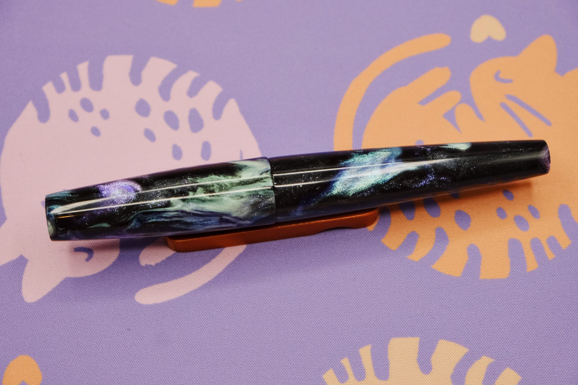 A torpedo-shaped fountain pen with flat ends made of black resin with purple shimmer, creamy aqua, and blue shimmer swirls, sitting on a wooden pen rest, with a purple deskmat with pink and orange curled up kitty pattern underneath