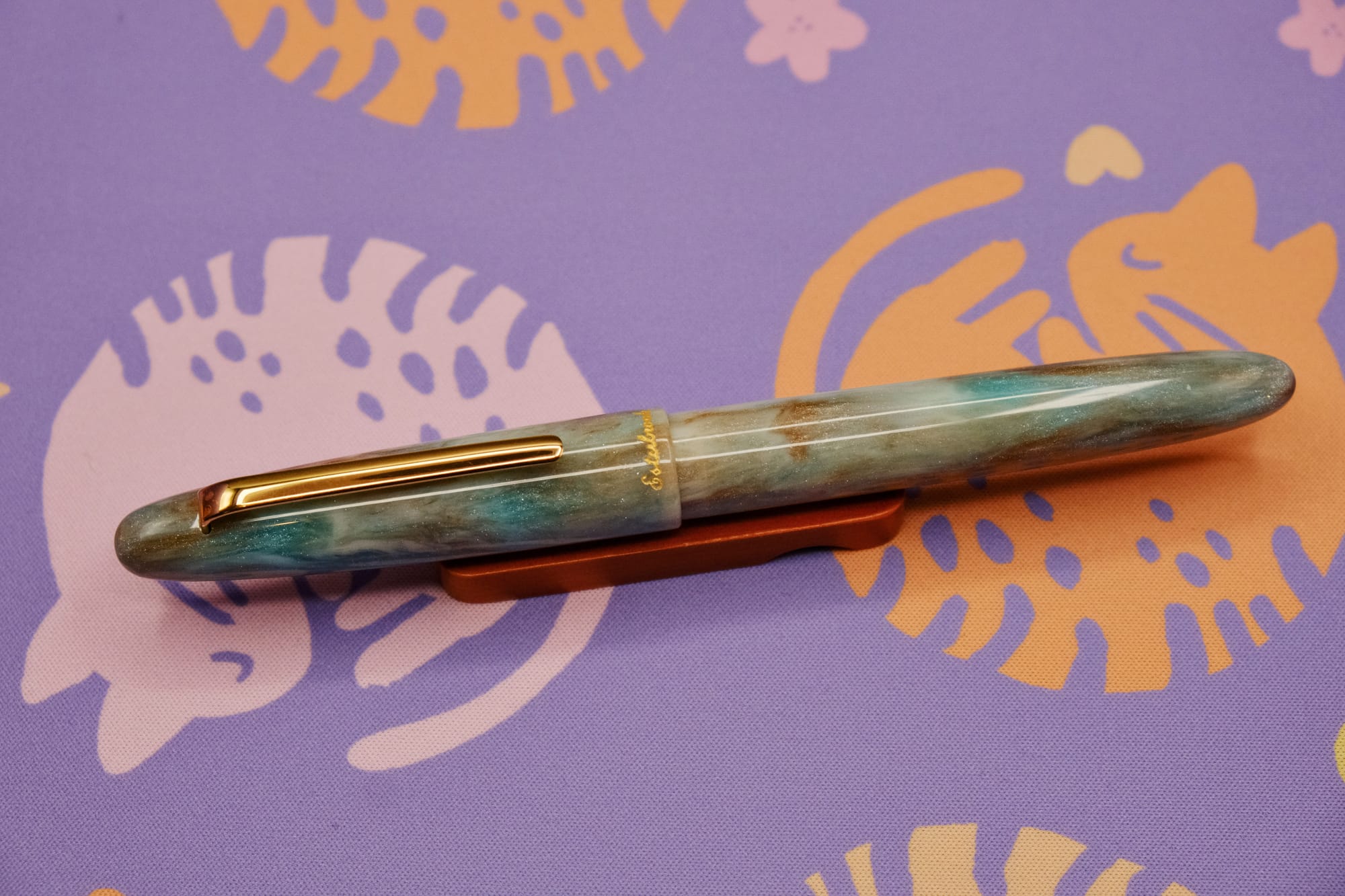 A cigar-shaped fountain pen with rounded ends and a gold clip, made of acrylic resin with bronze, blue-green, and creamy shimmer swirls, sitting on a wooden pen rest, with a purple deskmat with pink and orange curled up kitty pattern underneath