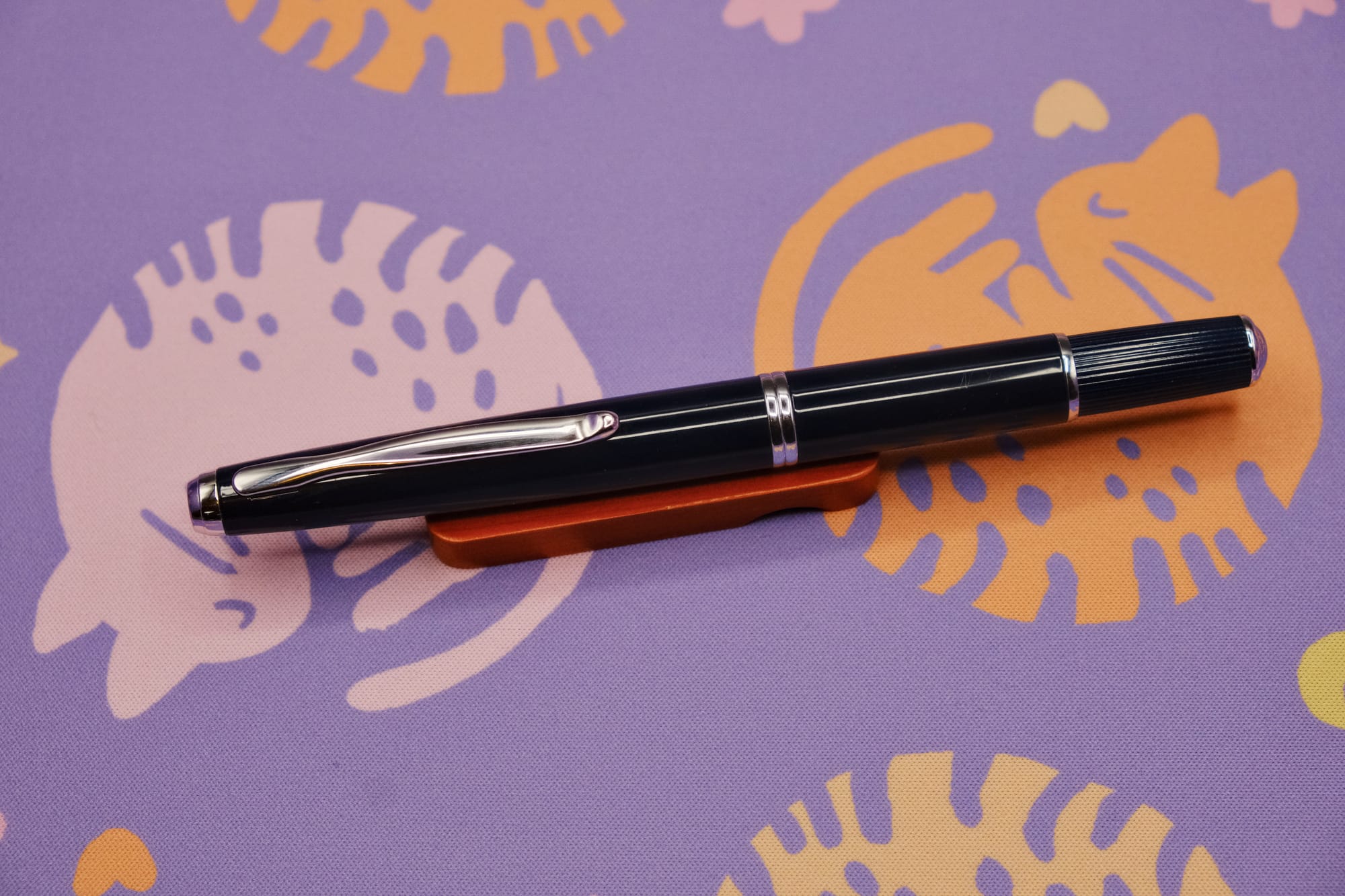 A very dark blue, retractable fountain pen with silver trim, sitting on a wooden pen rest, with a purple deskmat with pink and orange curled up kitty pattern underneath