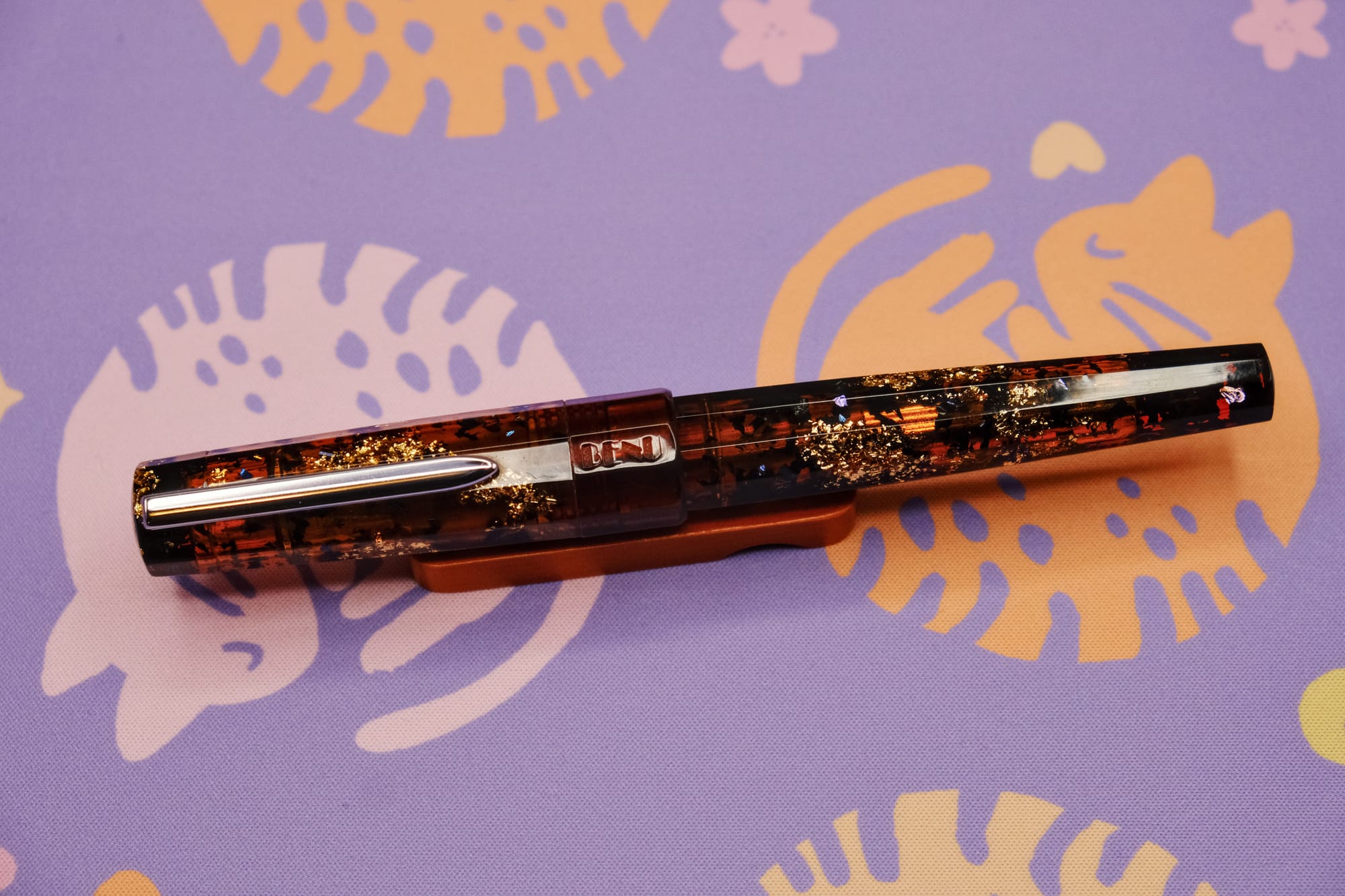 A large faceted fountain pen with a silver clip and rounded flat ends made of transparent tea brown resin infused with chunky gold and blue glitter, sitting on a wooden pen rest, with a purple deskmat with pink and orange curled up kitty pattern underneath