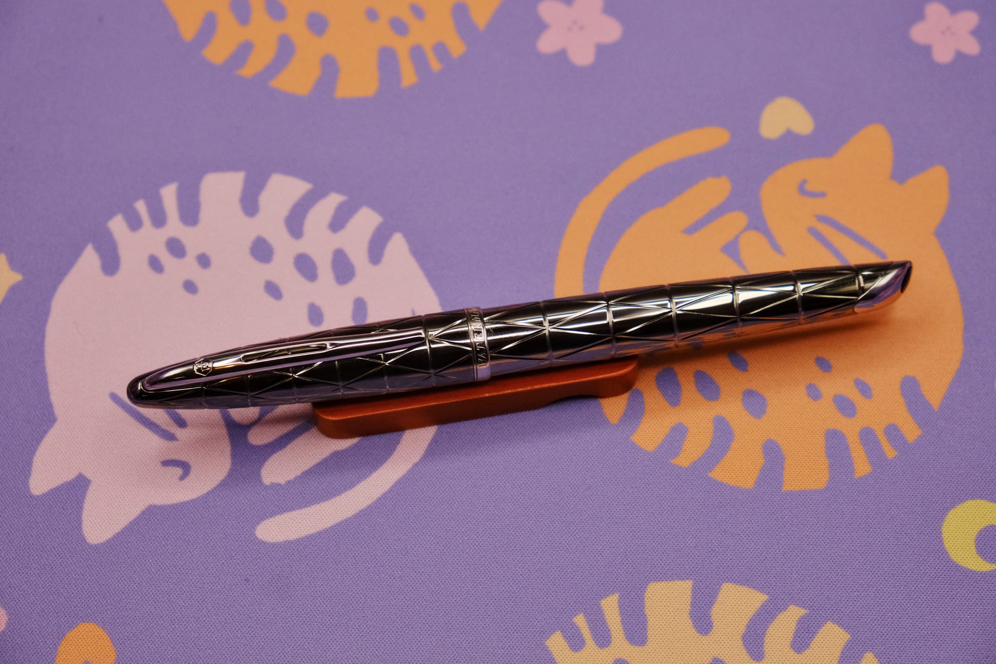 A medium-sized, torpedo-shaped, silver metal fountain pen with a geometric etched pattern, sitting on a wooden pen rest, with a purple deskmat with pink and orange curled up kitty pattern underneath