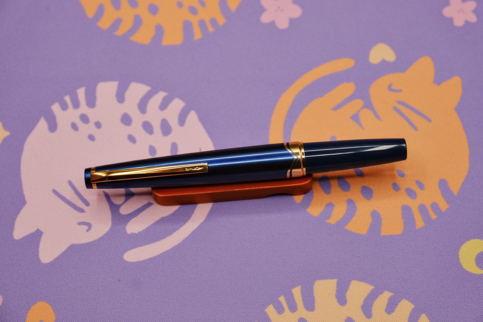 A metallic dark blue pocket fountain pen with gold trim and flat ends, sitting on a wooden pen rest, with a purple deskmat with pink and orange curled up kitty pattern underneath