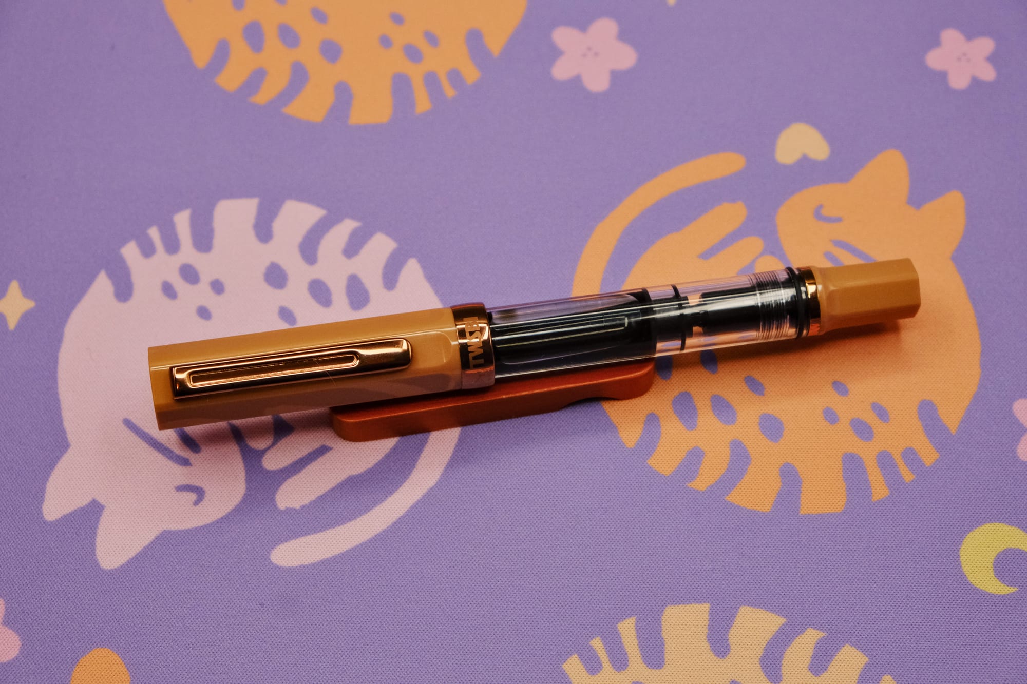 A clear-bodied demonstrator fountain pen with light coffee brown plastic cap and piston and bronze trim, sitting on a wooden pen rest, with a purple deskmat with pink and orange curled up kitty pattern underneath