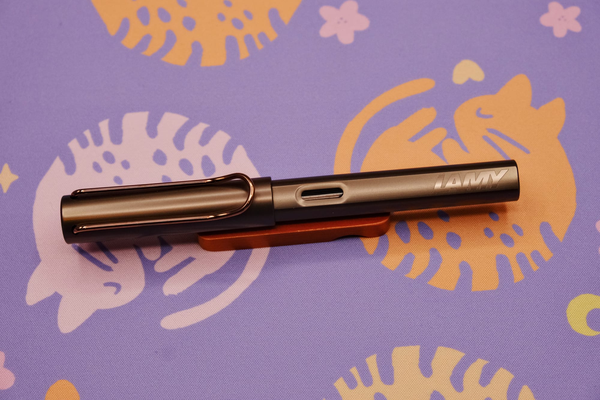 A metallic brown/bronze fountain pen with a bronze paper-clip-like clip, sitting on a wooden pen rest, with a purple deskmat with pink and orange curled up kitty pattern underneath