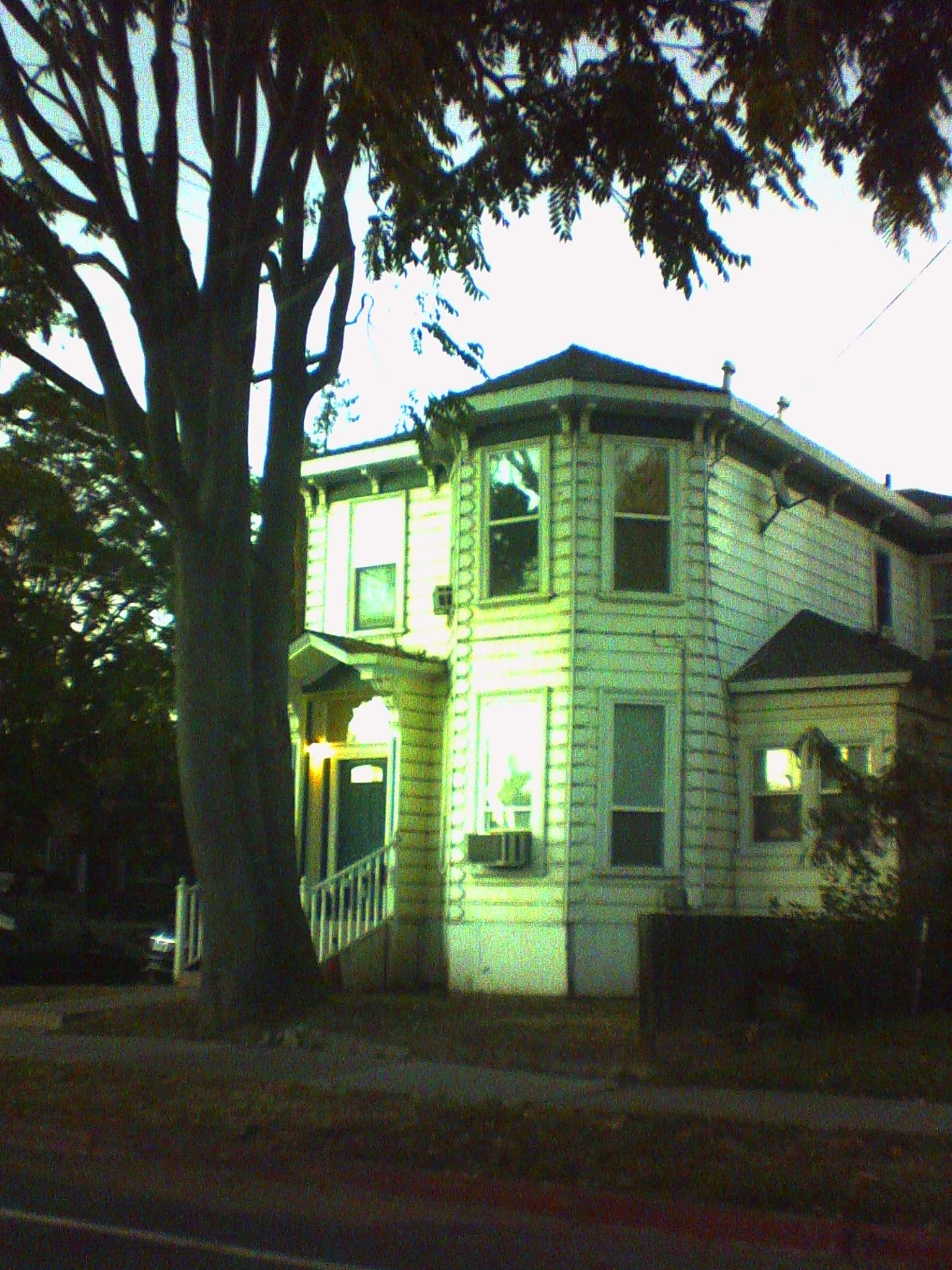 Low-res, green-tinged picture of an old white, 2-story house with a large tree right out front, its tall branches framing the top of the picture 