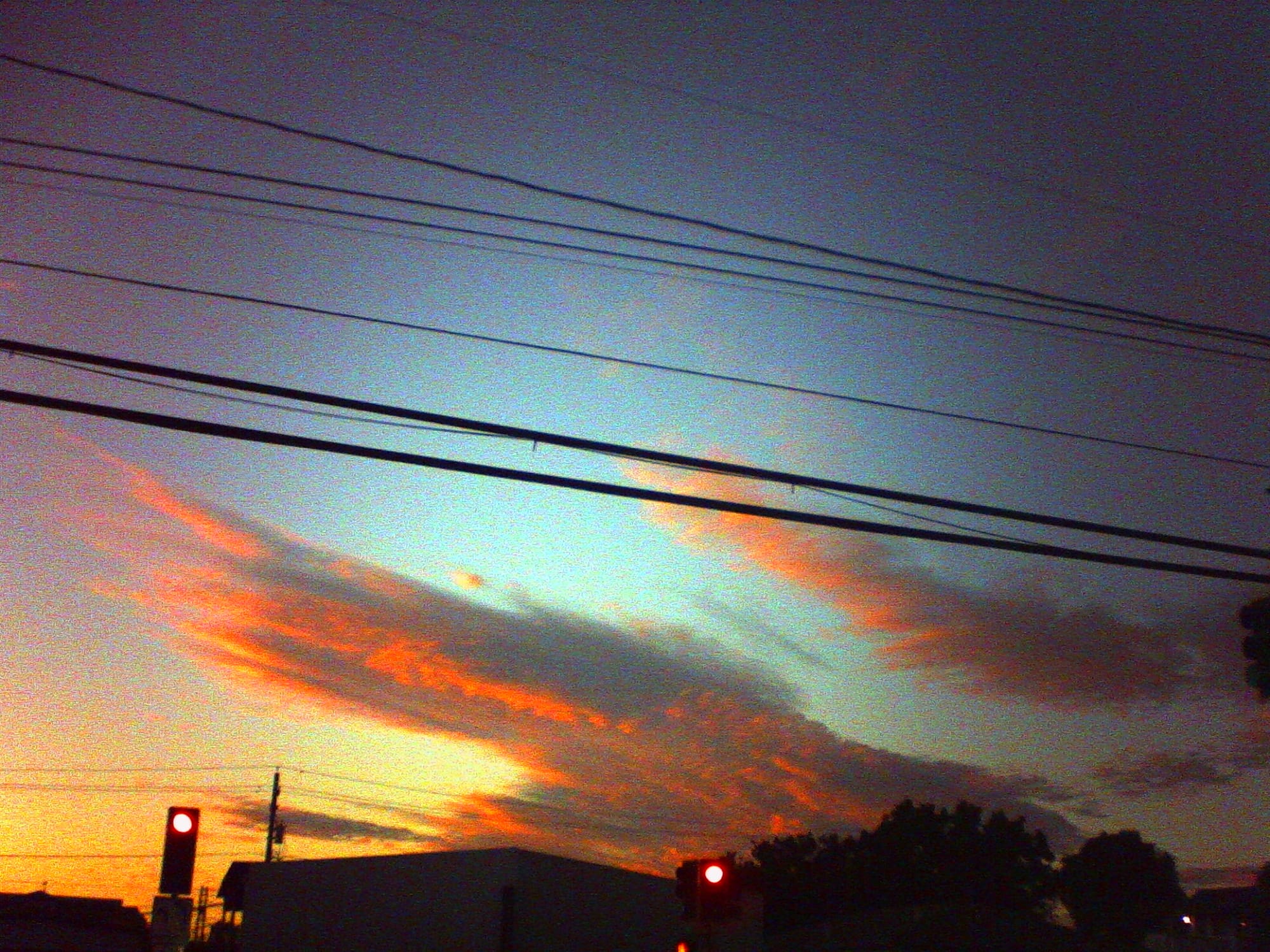 Low-res, noisy image with punchy colors of wispy layers of orange-red clouds in a blue-to-orange gradient sky, and traffic lights visible at bottom of the frame