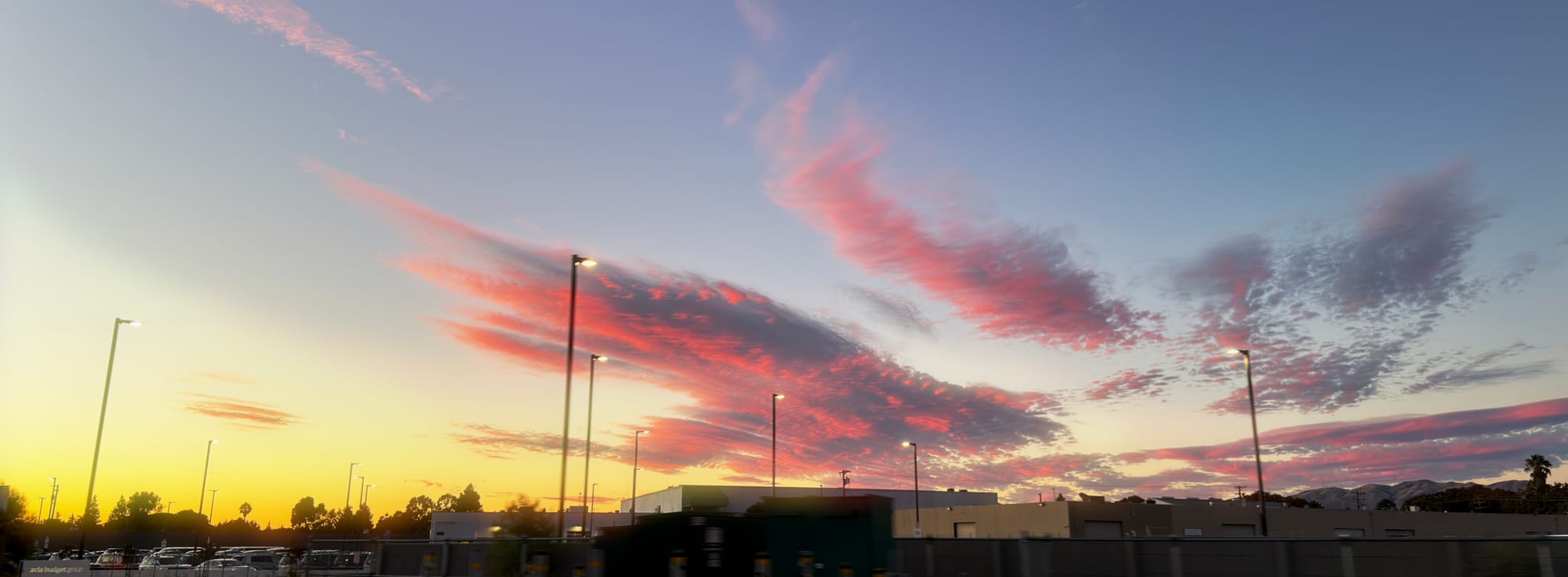 Wide-angle, panoramic shot of the blue-yellow-orange gradient sky with dark pink and gray swathes of clouds panning out from a spot on the horizon, a parking lot and tall lights in the foreground