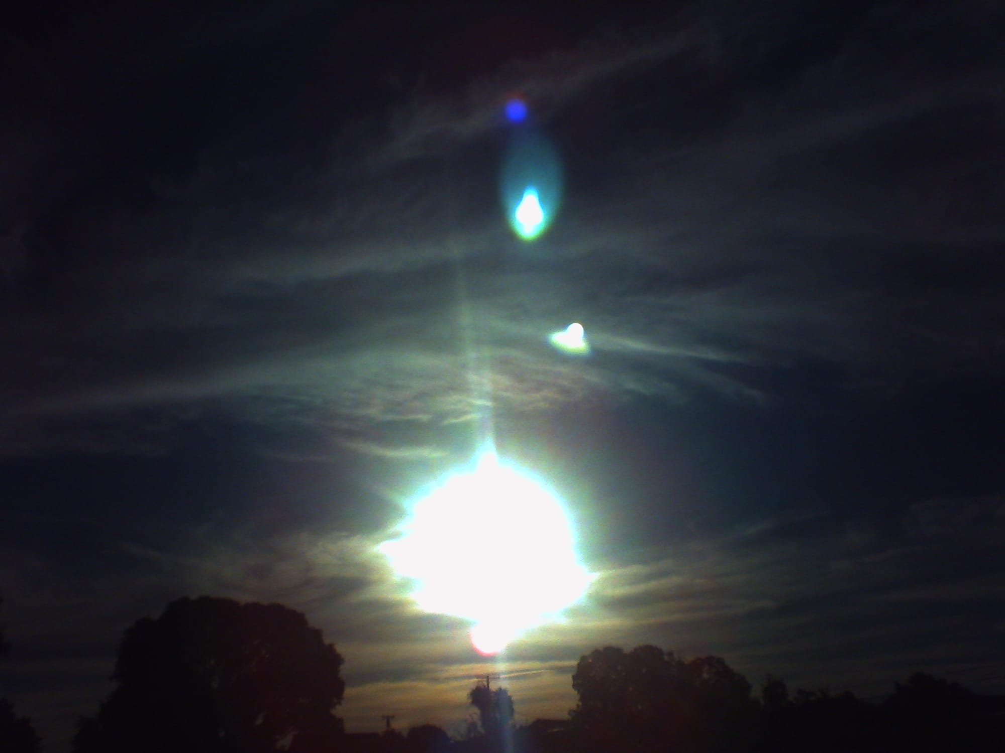 Shooting directly into the sun, underexposed partially cloudy sky surrounding the ball of light, lens flares at the top of the frame, treetop sihouettes along the bottom of the frame