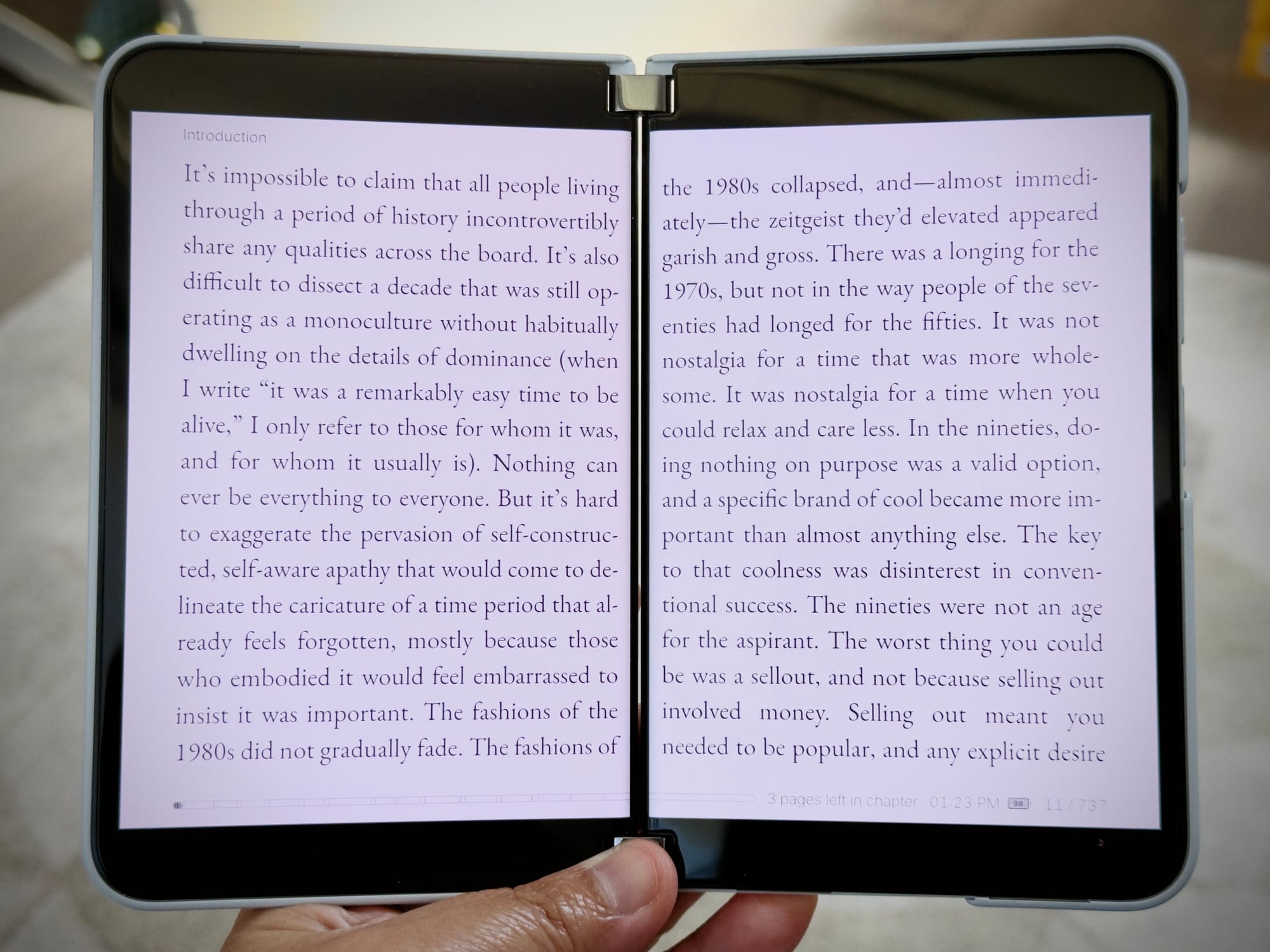 A hand is holding a Microsoft Surface Duo 2 open and showing e-book pages, with the hinge slightly bent, making the device look like a small book