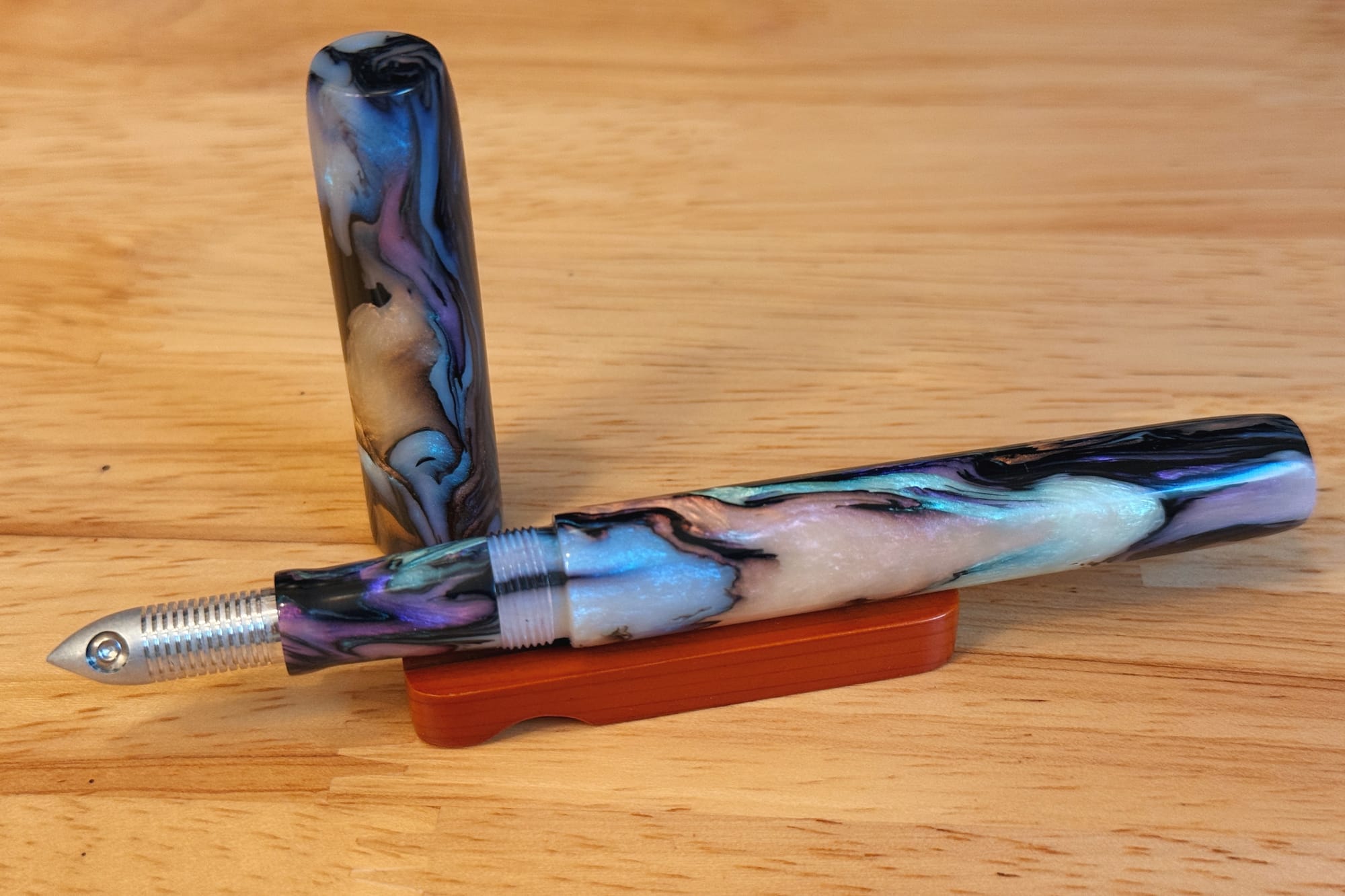 Swirly acrylic fountain pen with shimmery black, opal, peach, purple, blue, and pink colors fitted with the aluminum L'il Dipper nib, pen cap sitting upright behind the pen body