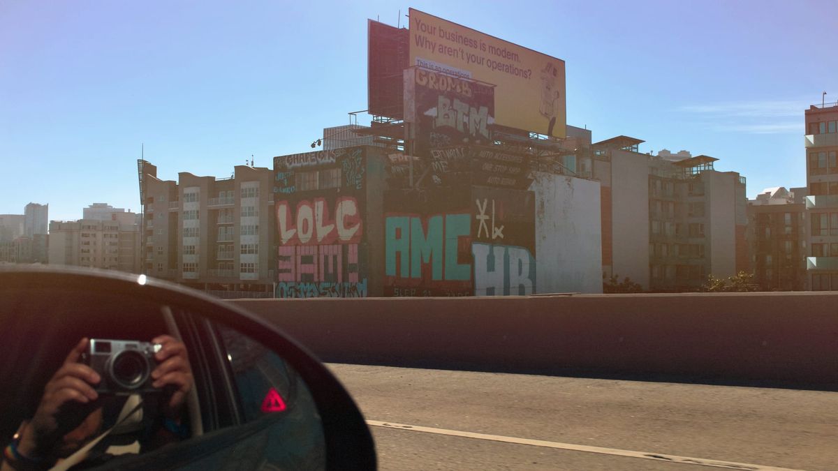 Image shot out the passenger window of a graffitied building along Hwy 101 in SF, photographer reflected in side-view mirror