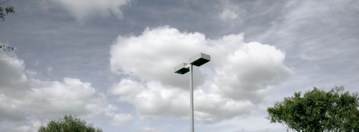 Panorama of a sunny, party cloudy sky, with a parking lot lamp in the middle of the shot, obscuring a poofy cloud behind it, tree tops scattered around the left and bottom of the frame