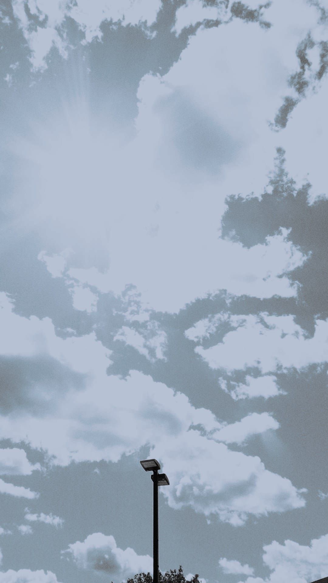 Vertical 16:9 image looking directly at the sun and a partly cloudy sky, a parking lot lamppost jutting up from bottom edge, disrupting sky