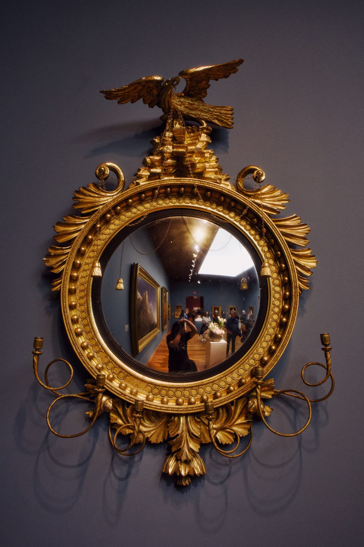 An old, golden, ornate fisheye mirror hanging on a blue-gray wall in a museum, the photographer visible in the middle of the mirror