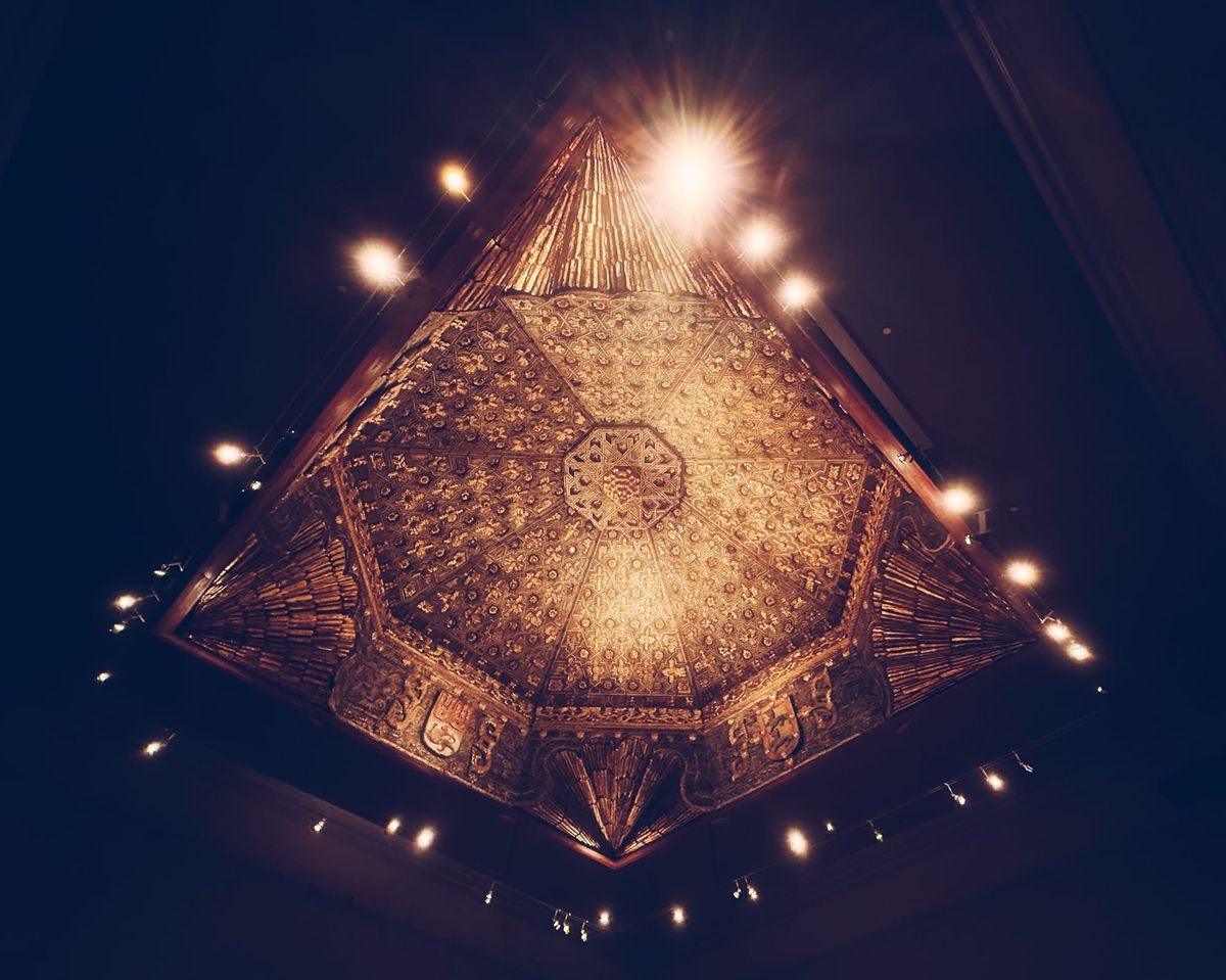 Looking up at an ornate, bronzy ceiling with spotlights shining down around its edges, its shape distorted by the extra wide angle of the camera