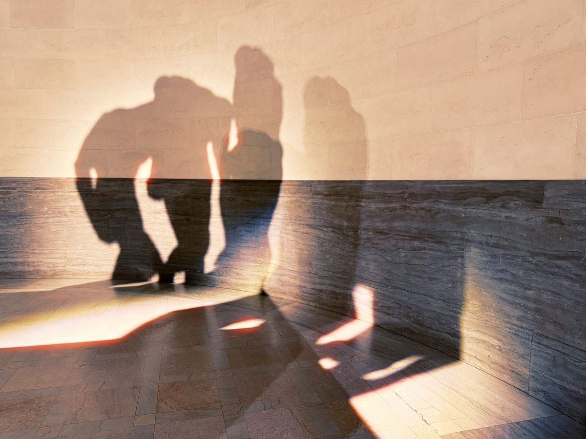 Abstract overlapping sculpture shadows of 3 individuals cast against a two-toned, slightly curved museum wall, ivory at top, dark gray bottom half