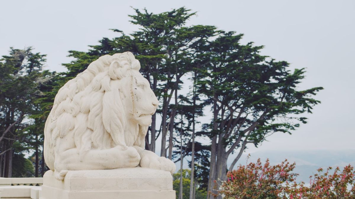 A stone sculpture of a lion with tall cypress (?) trees in the background, a hazy horizon visible beyond the trees