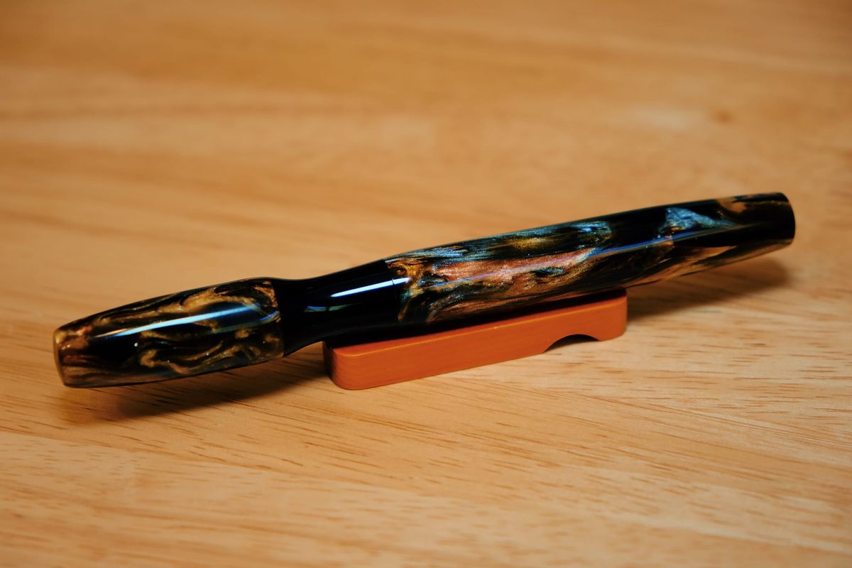 A fountain pen made out of a dark resin with swirls of golden brown and silver shimmer, sitting on a small wooden pen rest