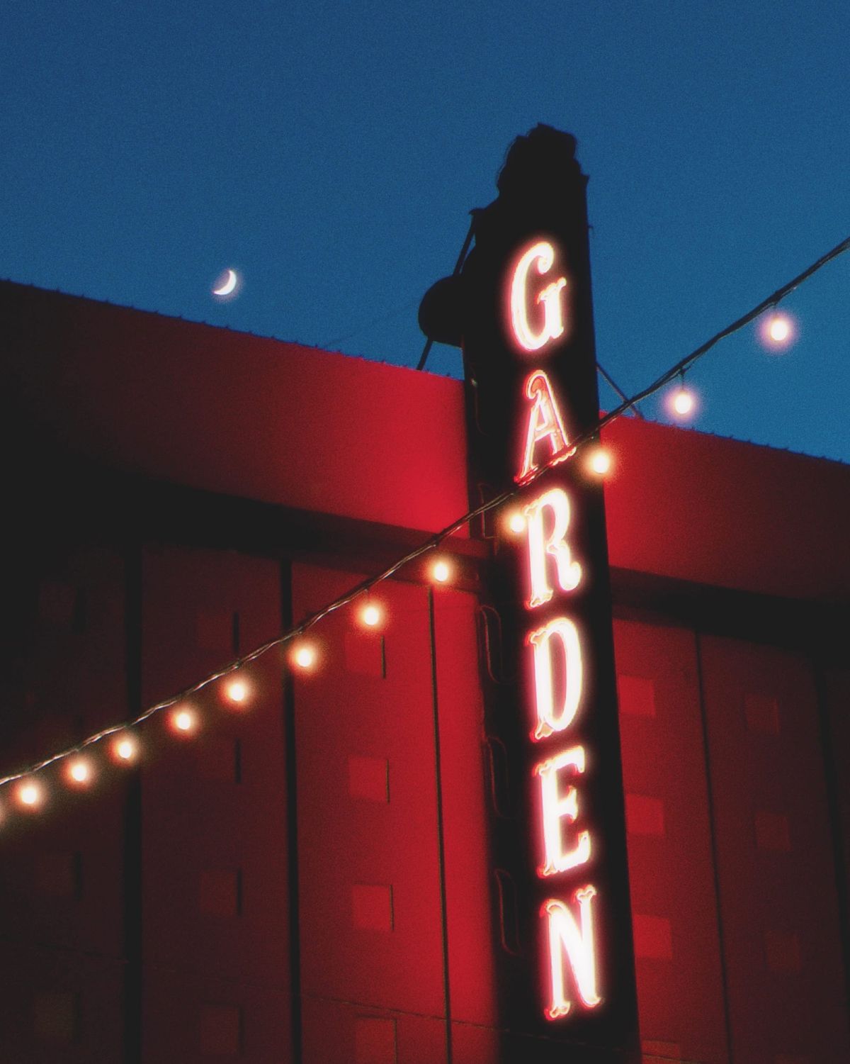 Dreamy image of a vertical neon sign that says "garden", a string of white lights across the foreground, and a crescent moon in the background