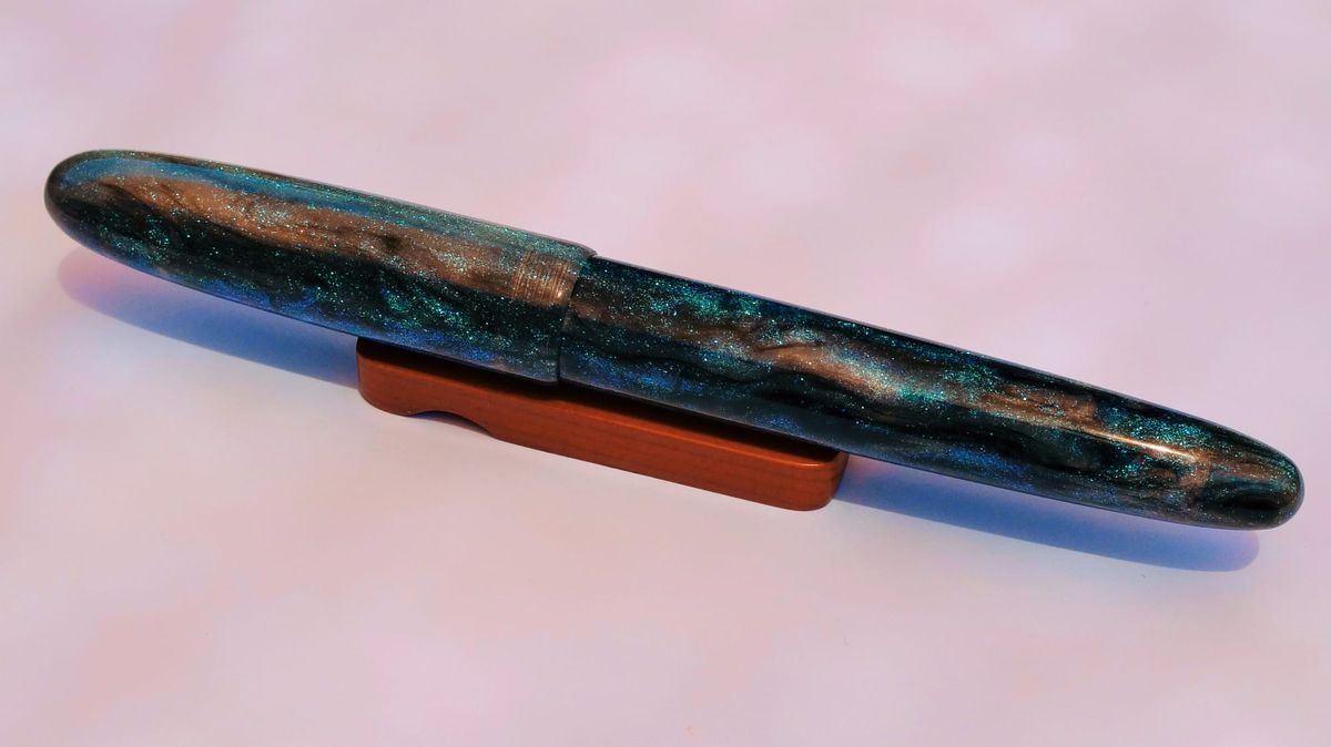 A glittery, cigar-shaped fountain pen with dark, translucent resin and light blue glitter sitting on a small, reddish wooden pen rest