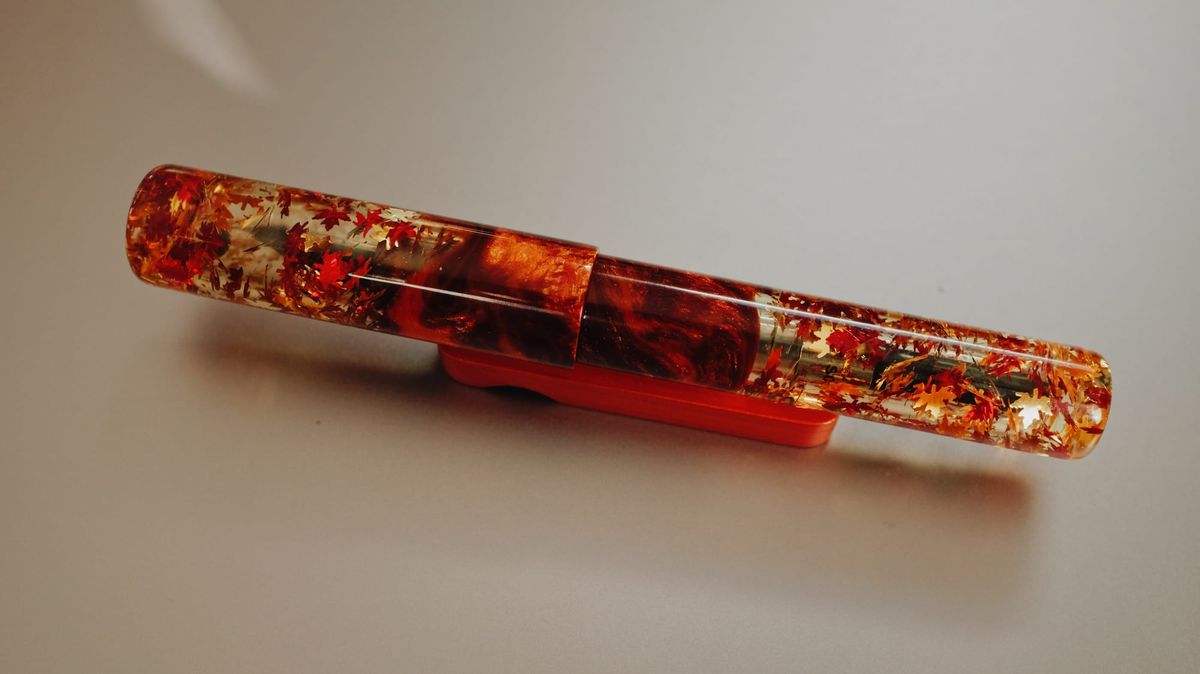A golden brown and red, fall-themed resin fountain pen with chunky fall leaves glitter embedded in transparent resin at top and bottom of pen body, with faux burl resin in middle of pen body