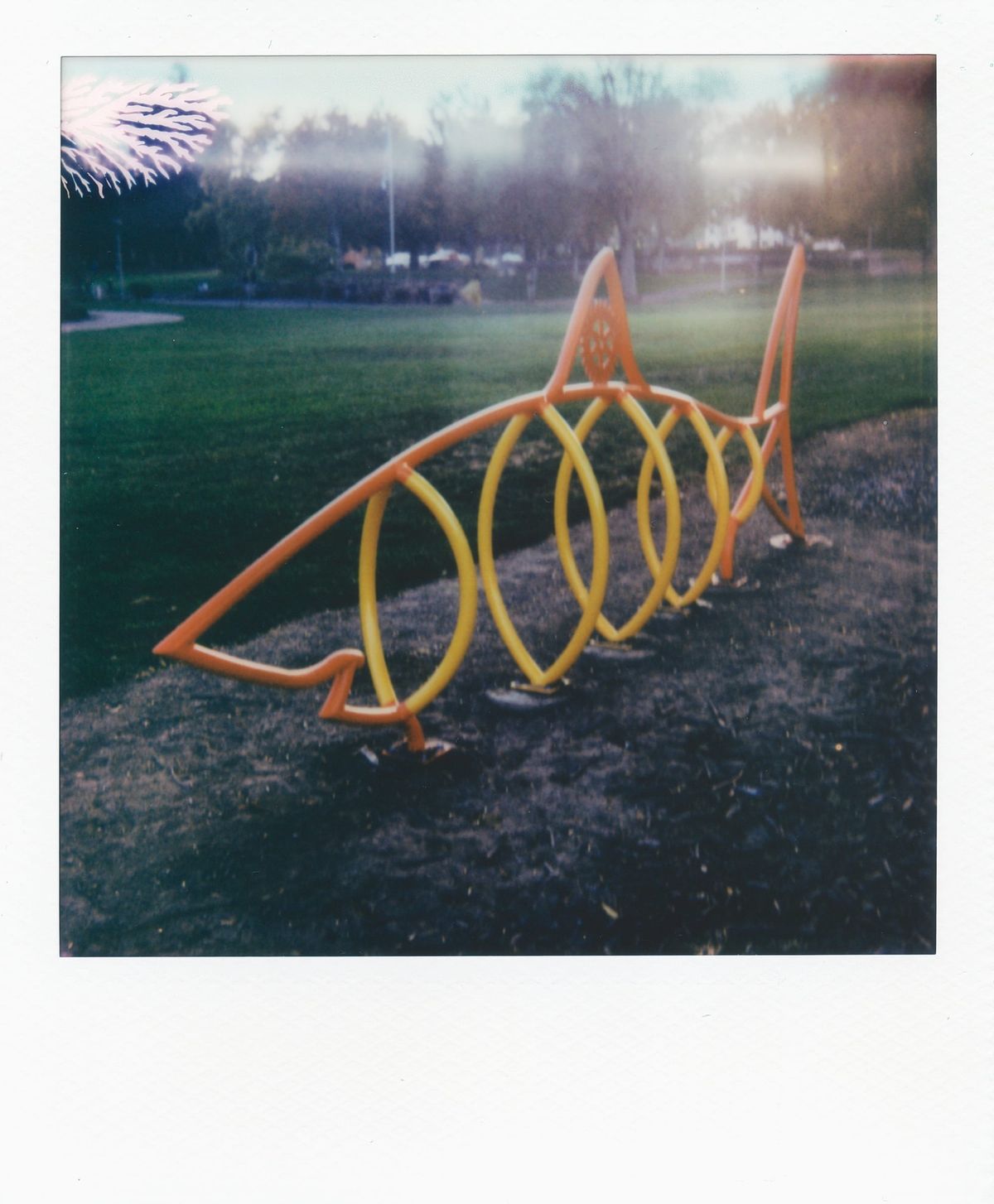 Lo-fi Polaroid picture of an orange and yellow shark-shaped playground apparatus, some random film development errors visible at the top of the frame