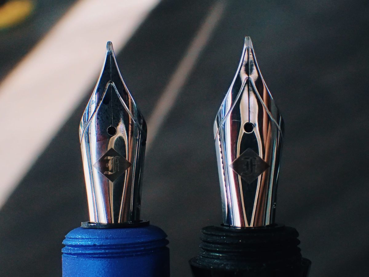 Close-up of two silver fountain pen nibs, the fine one mounted in a blue section, the medium one mounted in a black section