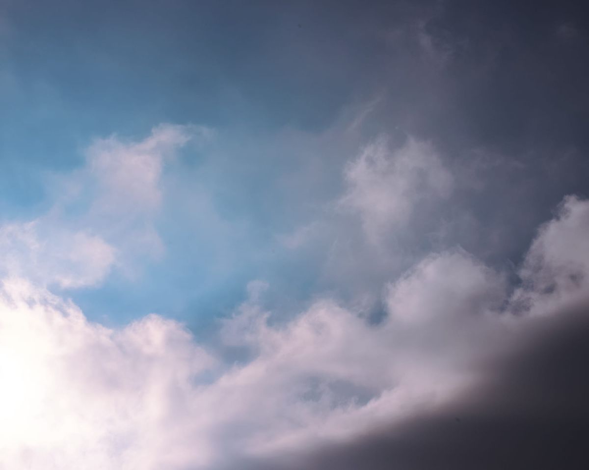 Painterly, dramatically lit clouds in a medium blue sky, lower left showing strong sunlight, lower right corner showing dark shadows