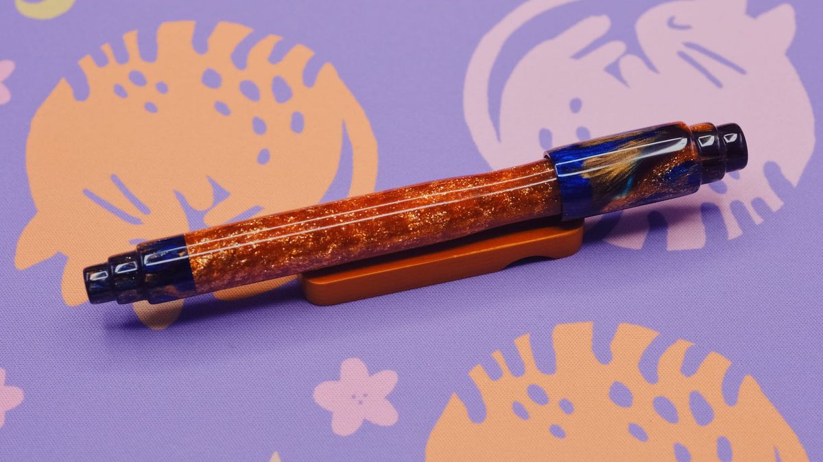 A very sparkly pen with unusual shaped cap and end finial sitting on wooden pen rest. The middle of the pen body is bright shimmery orange, while ends are dark blue with shimmery swirls.