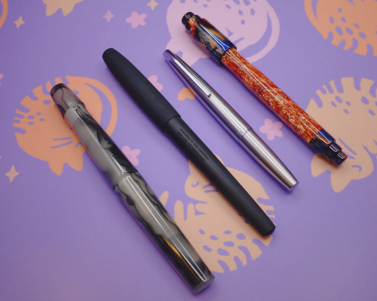 4 fountain pens sitting on purple deskmat: gray and black swirly acrylic, black ebonite, stainless steel pocket pen, and vivid copper shimmer with blue, gold, peacock swirly acrylic end caps