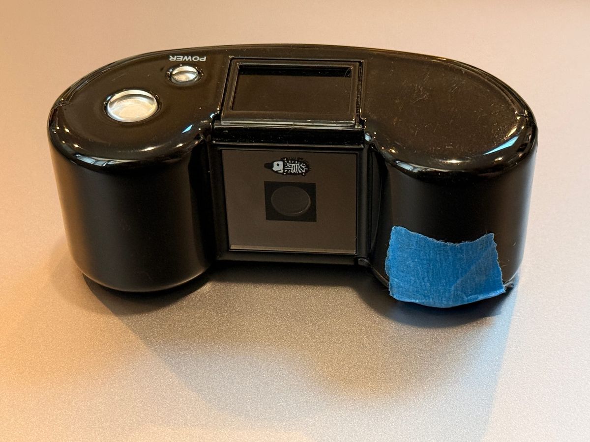 Close up of a small, black digital camera, similar in shape to a roll of 110 film, with a piece of blue tape seen on its lower front corner