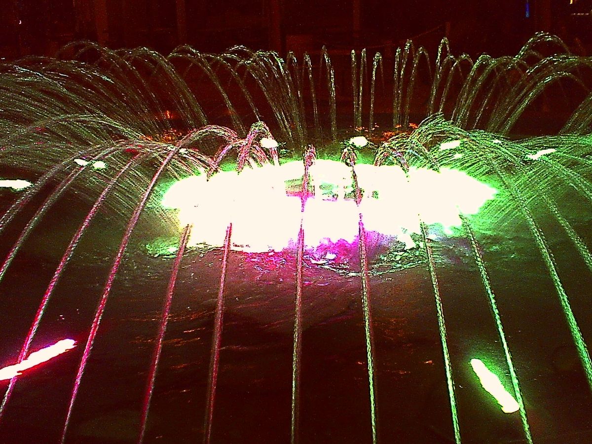 A very low-res, ugly, overexposed image of a fountain at the mall with bright lights in the center and many arcs of water flowing out around the center