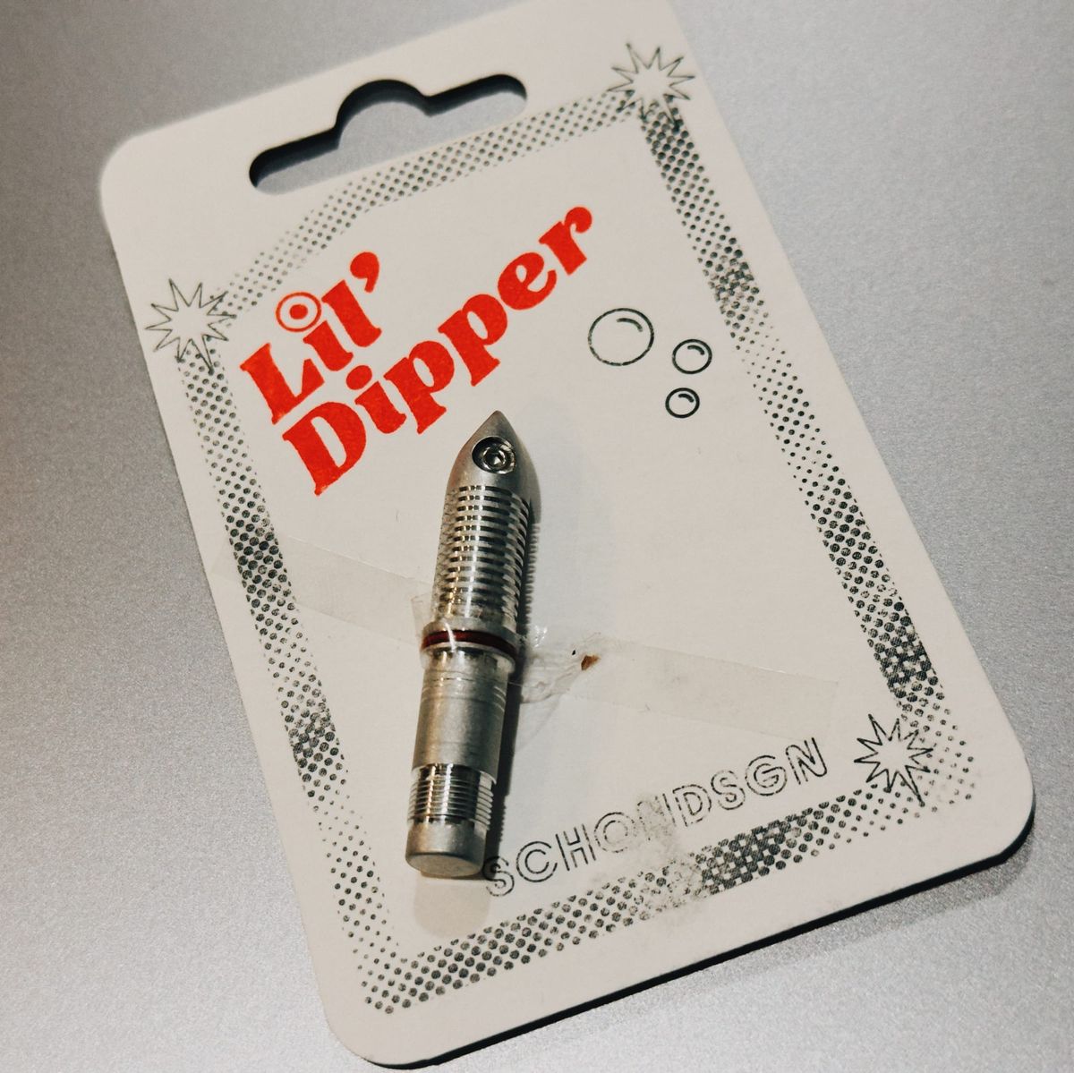 An aluminum, bullet-shaped dip nib with a small screw near the top and horizontal ridges beneath, taped to a paper card backing labeled L'il Dipper at the top, and SCHONDSGN at bottom