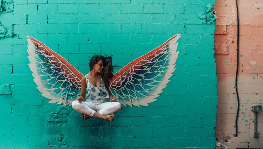 A woman appears to levitate, cross-legged, against a wall. Painted wings on the wall seem to sprout from her back.