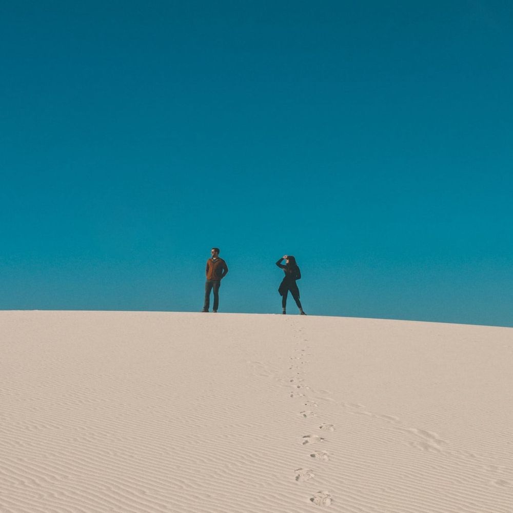 Two people standing in the middle distance on a sand dune, against the sky. They are looking intently at something out of frame.