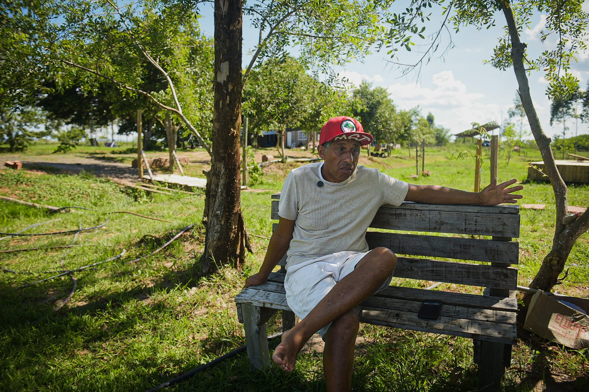 Homem sentado em banco de madeira em área rural arborizada, gesticulando durante conversa ao ar livre.