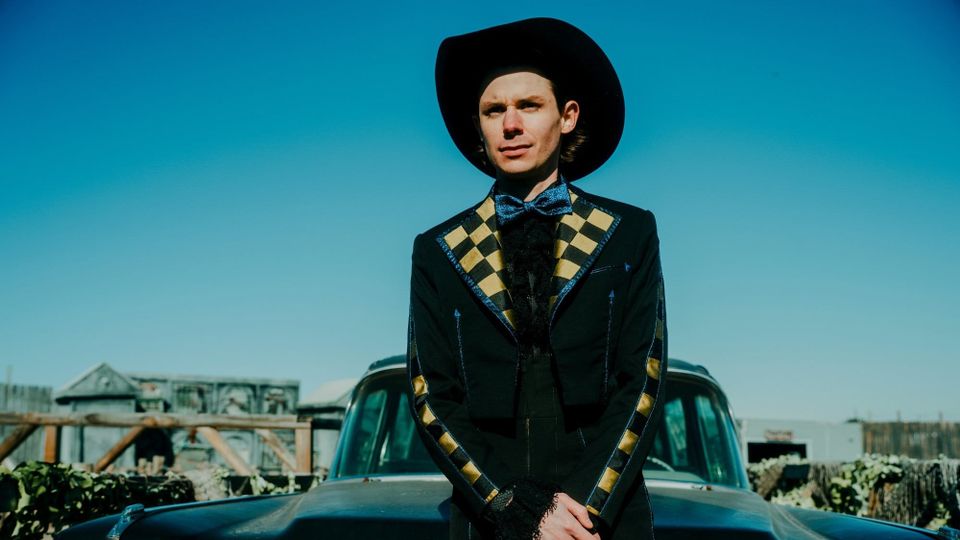 A photograph of Hayden Pedigo standing in front of a car wearing a suit and hat.