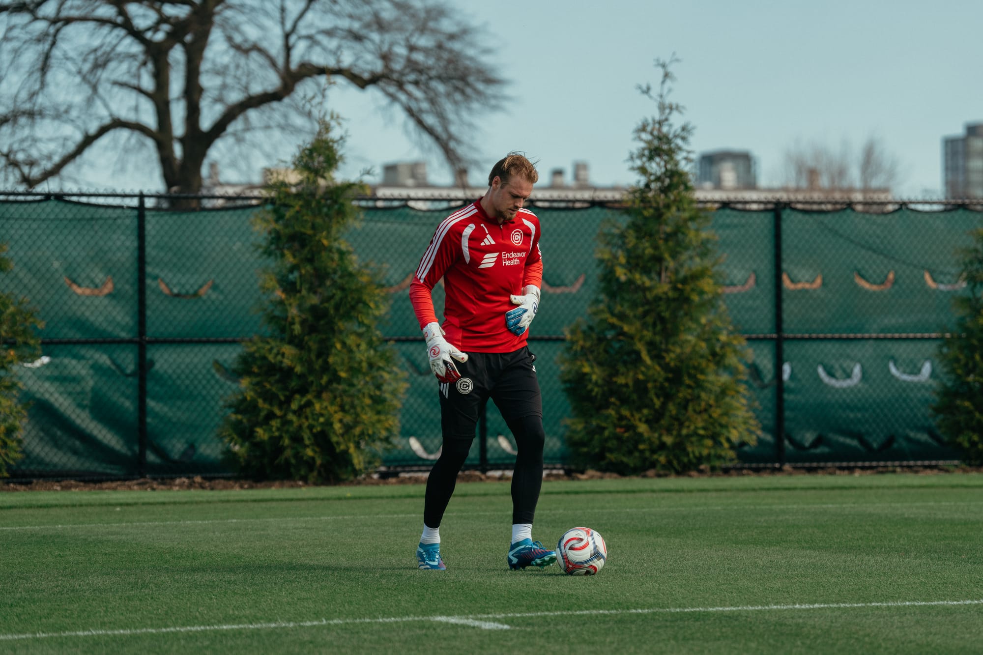 Chris Brady kicks the ball at training with shrubs and a fence in the background.