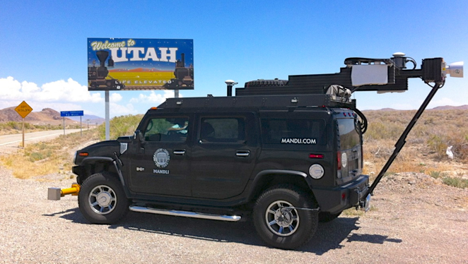 A color daytime photo of a black Mandli SUV with camera and scanning gear attached, on the side of a road next to a sign that says Welcome to UTAH.