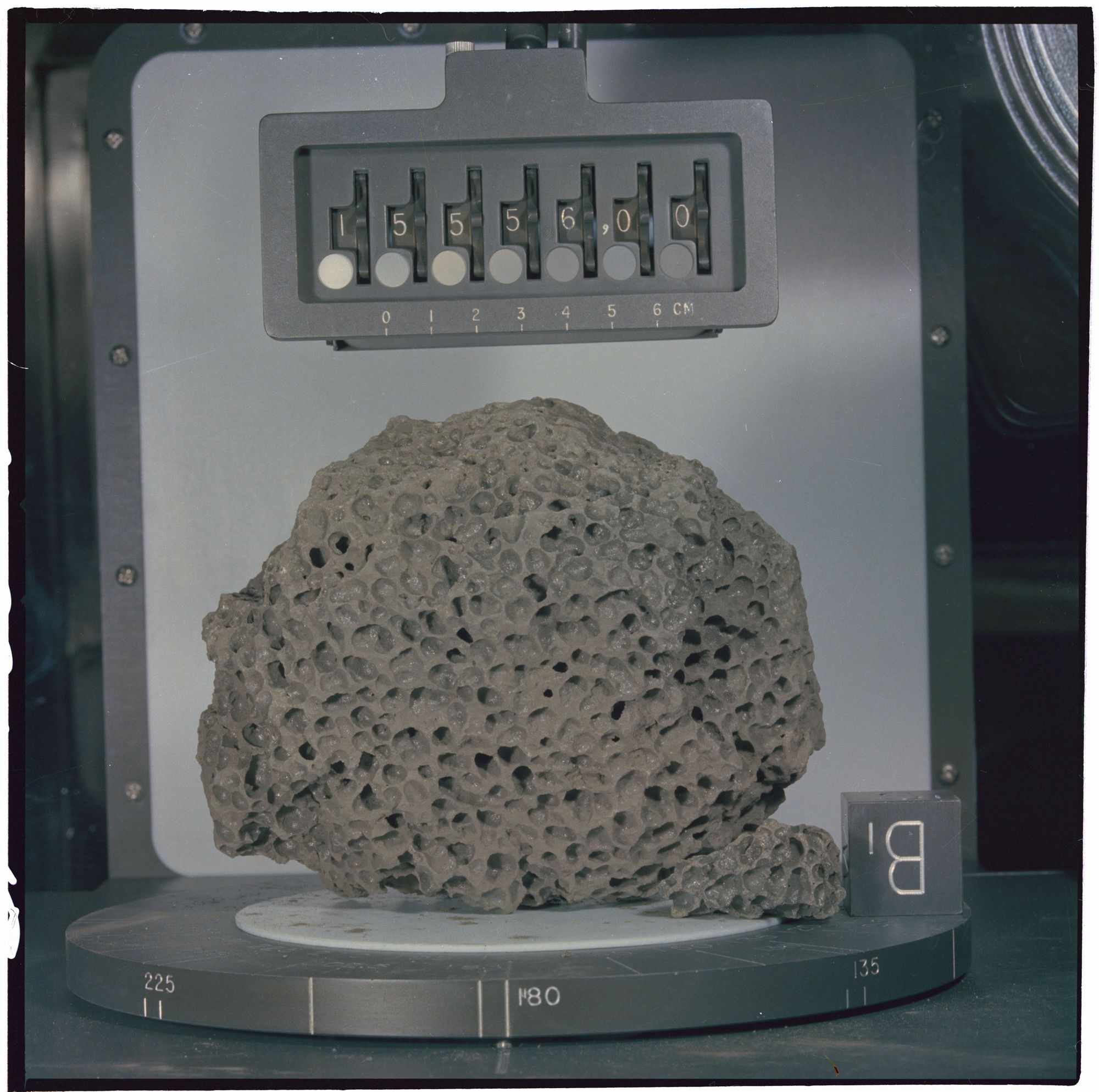 A photo of a pockmarked dark grey moon rock sitting on a circular metal surface with calibration targets and numbers,