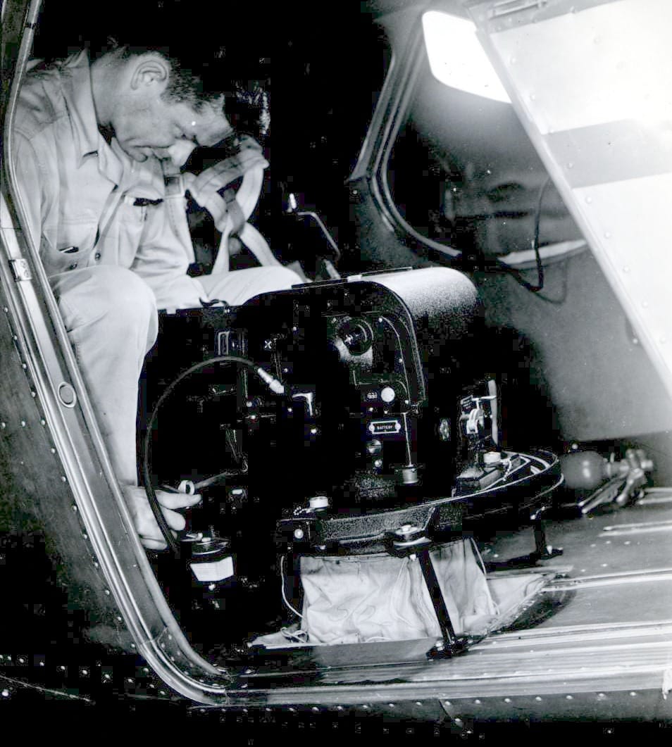 A black and white photo of a man looking down at a large black aerial K-17 camera mounted to the floor of a small airplane.