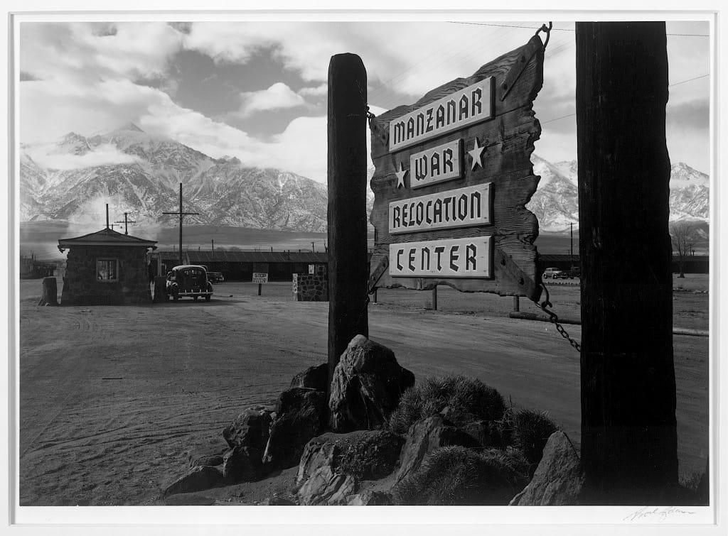 A black and white photo of a wooden sign that reads "MANZANAR WAR RELOCATION CENTER" against a mountain range in the distance. 