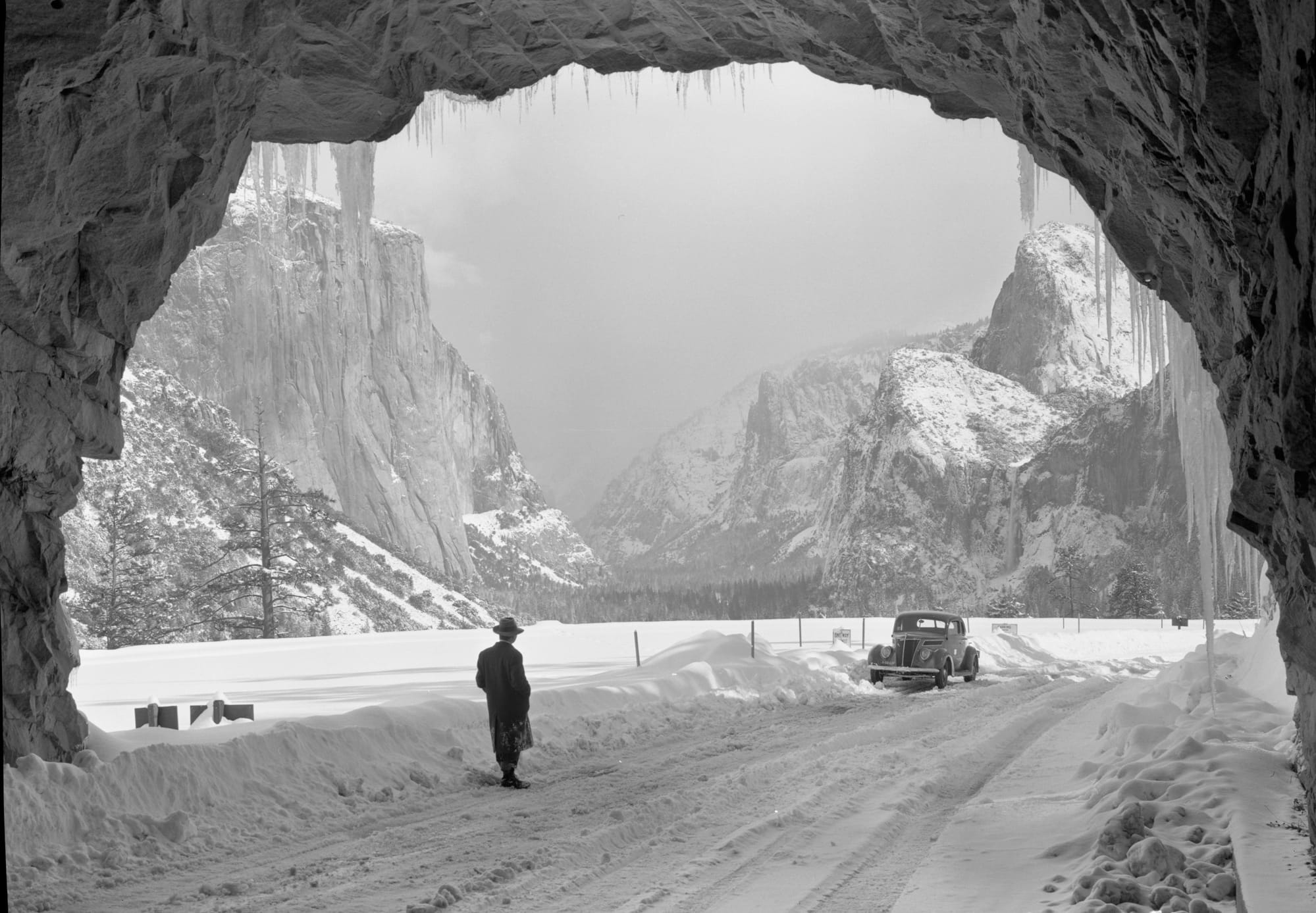 A black and white photo of a snowy scene of Yosemite valley from inside a tunnel with a man and an old car. 