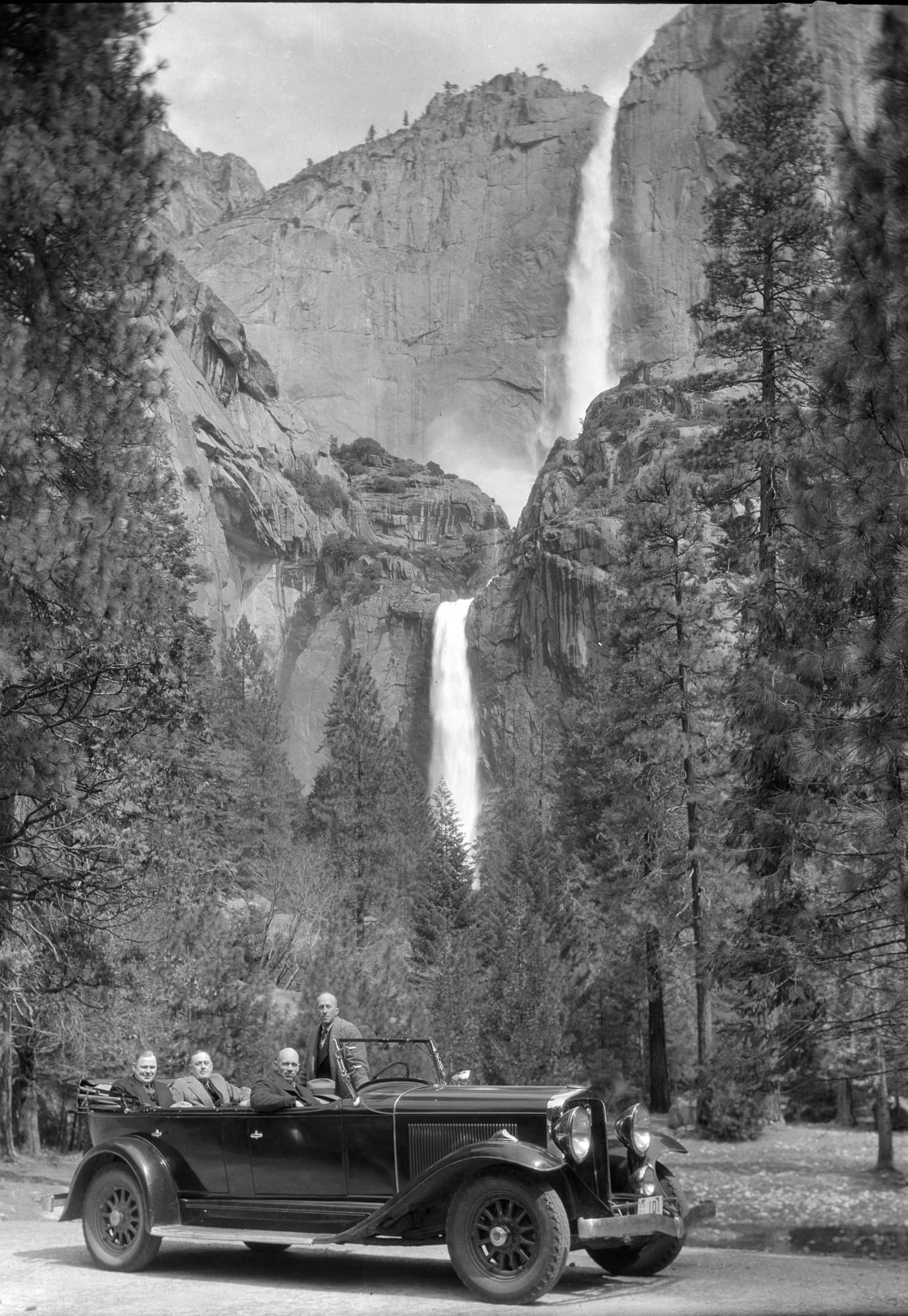 A black and white photo of an old car with four men in front of two tall waterfalls in Yosemite. 