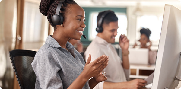 Smiling call center employee claps while speaking into a headset, with coworkers visible in the background, suggesting a positive and collaborative atmosphere