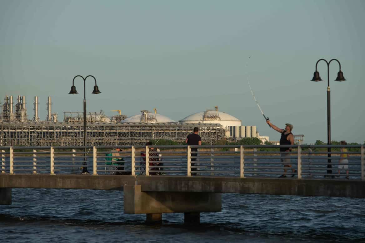 People fishing on a pier with an LNG terminal seen in the distance