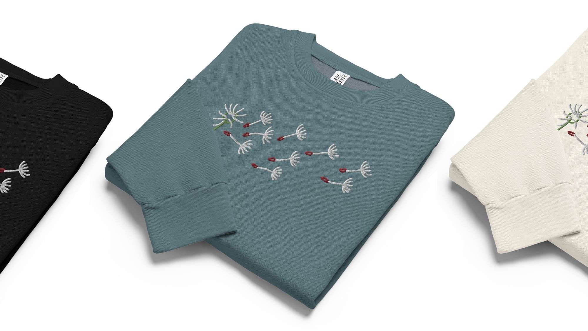 Folded crewneck sweaters featuring a clean embroidered dandelion with wind-blown seeds.
