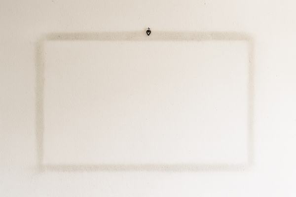 A plain off-white wall showing a faint rectangular outline where a picture frame once hung, with a single metal hook still in place.