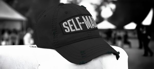 A close-up of a Self-Made Distress Dad Hat resting on a white surface with figures behind it at a public event.  